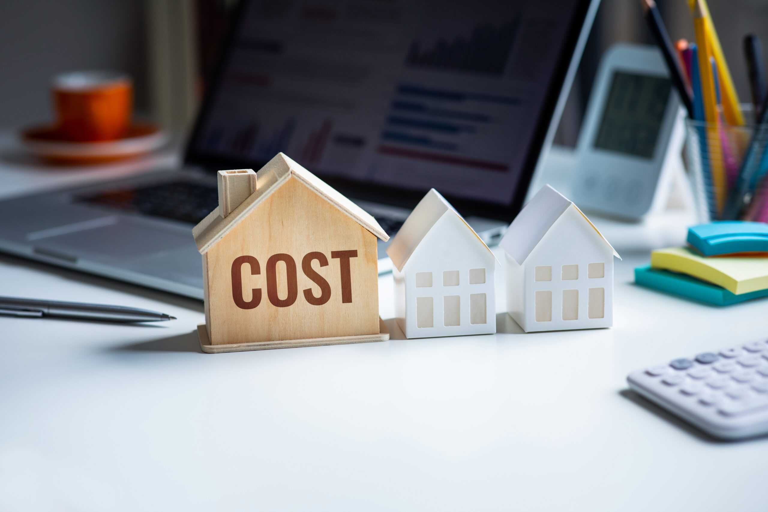 Three model houses on a desk, with one marked "COST" in front of a laptop displaying graphs. Nearby are pens, pencils, a calculator, a clock, and an orange cup.