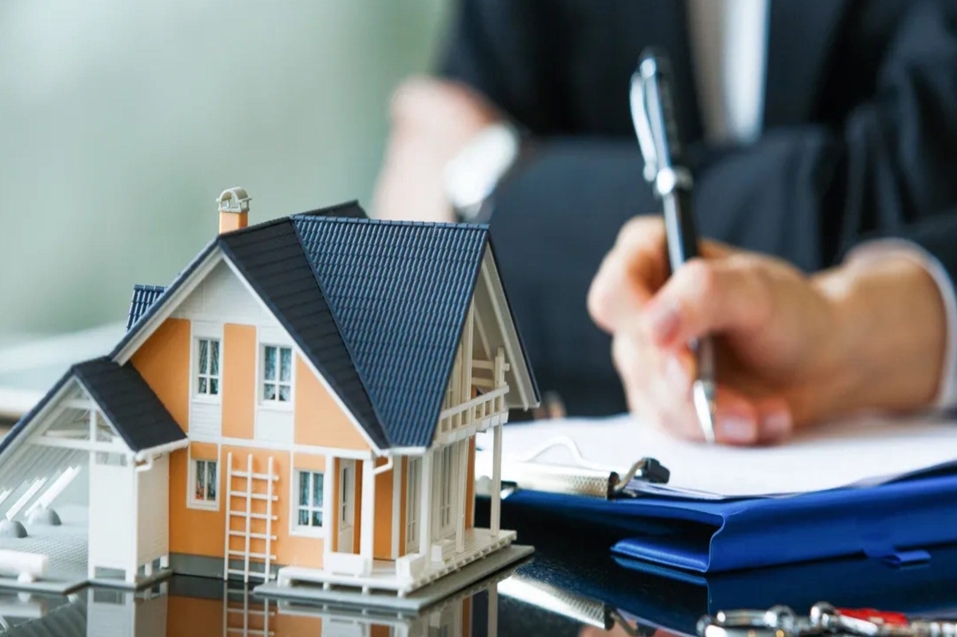 A model house sits on a desk next to documents and a clipboard. A person in a suit is signing papers in the background.
