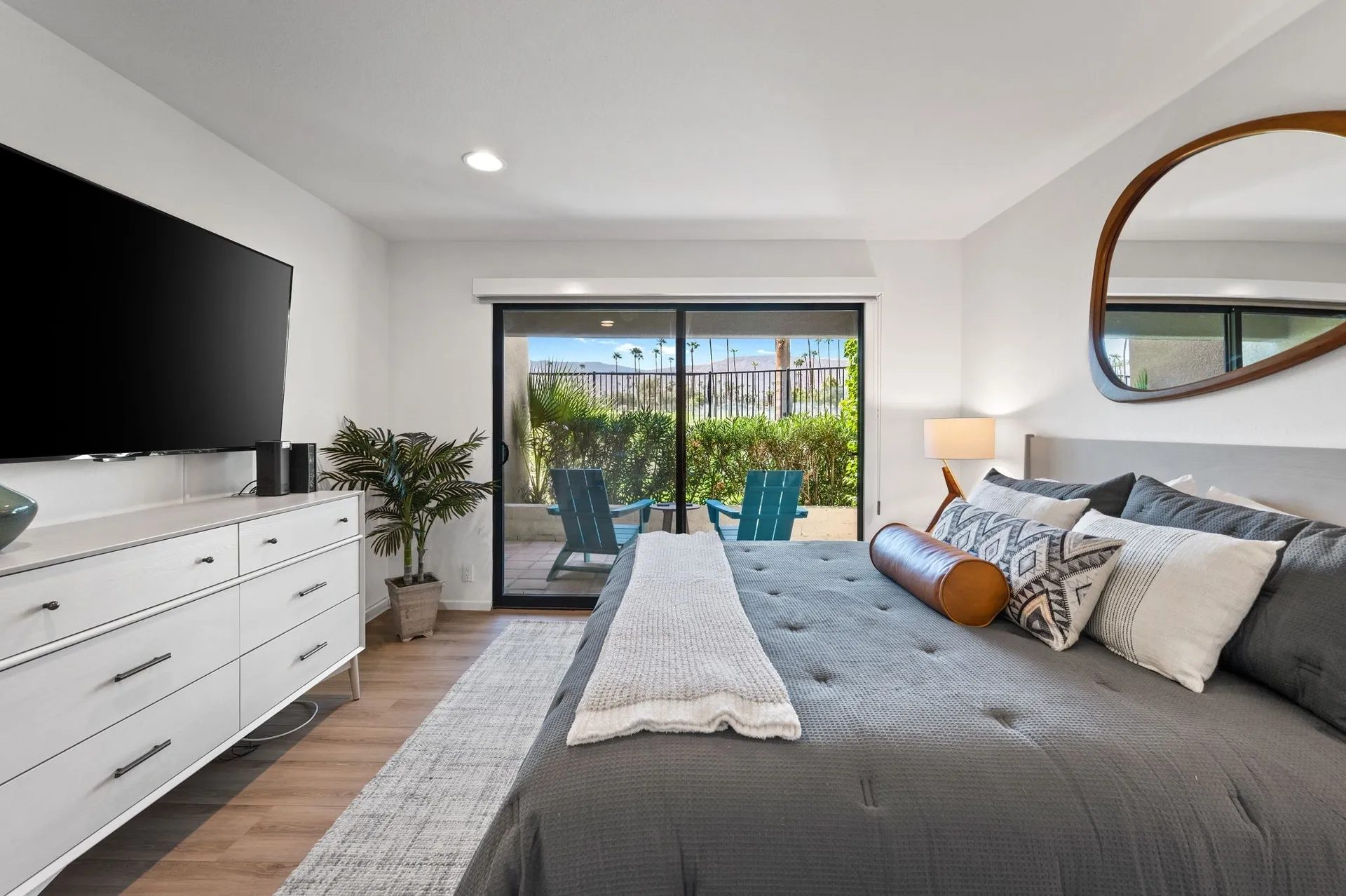 A modern bedroom featuring a gray bedspread, decorative pillows, a large mirror, a flat-screen TV on a white dresser, and a sliding glass door opening to a patio with chairs and plants outside.