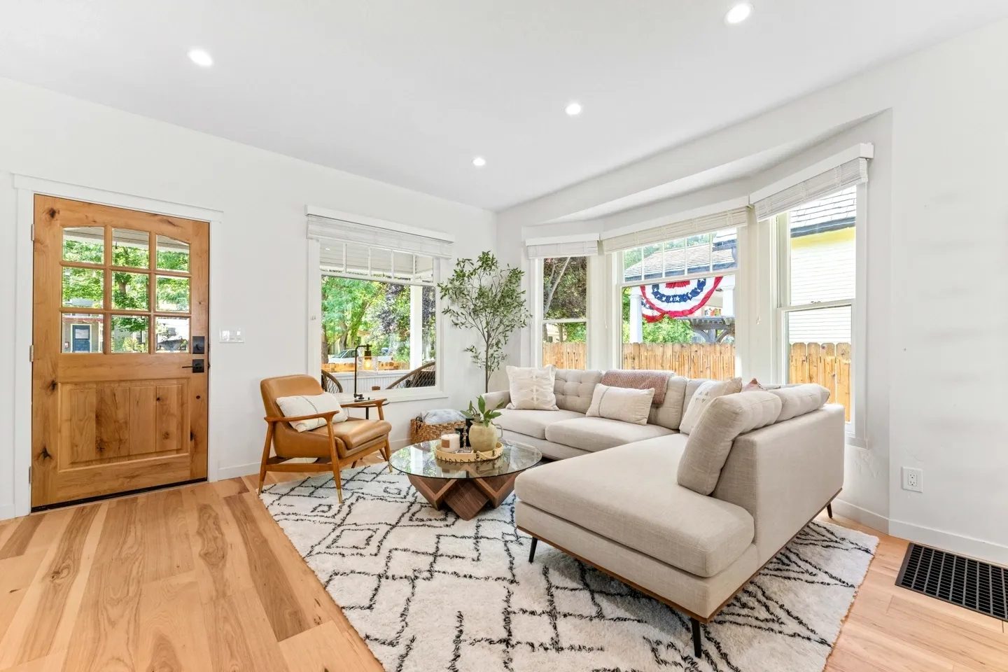 Living room with light wood floors, beige sectional sofa, glass coffee table, wooden armchair, and green plant near large windows with blinds. Wooden door on the left. Wall decor visible outside.