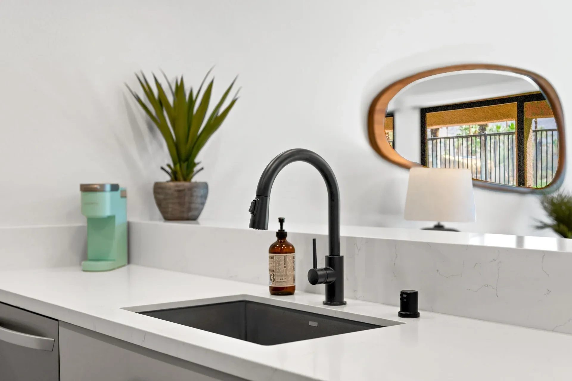 Modern kitchen sink area with a black faucet, white countertop, soap dispenser, potted plant, and a turquoise appliance. A mirror and table lamp are reflected in the background.