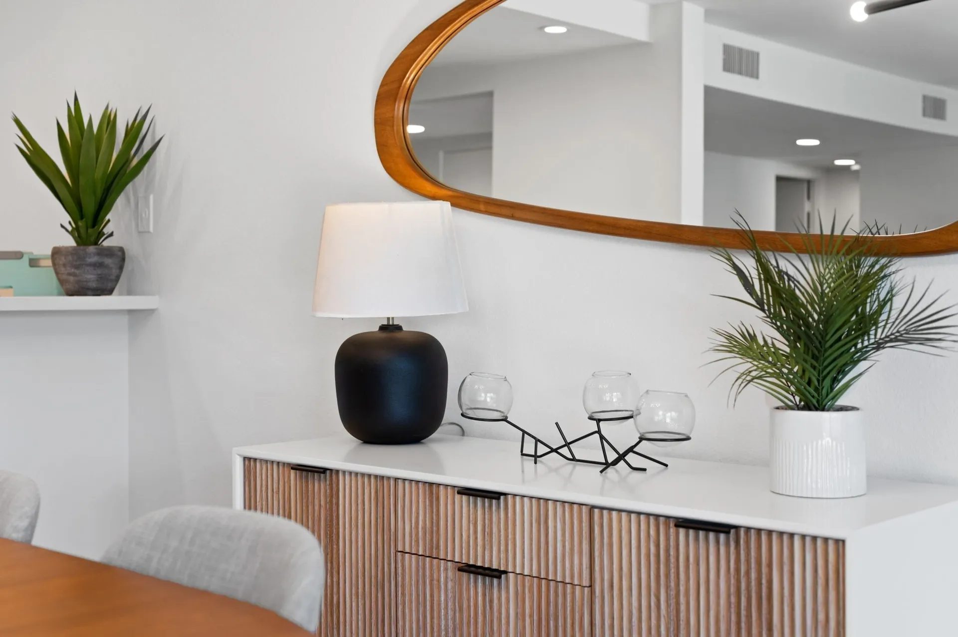 A modern interior showing a white sideboard with a wooden front, topped with a black lamp, three candle holders, and a plant. A large oval mirror hangs above, with a white wall in the background.