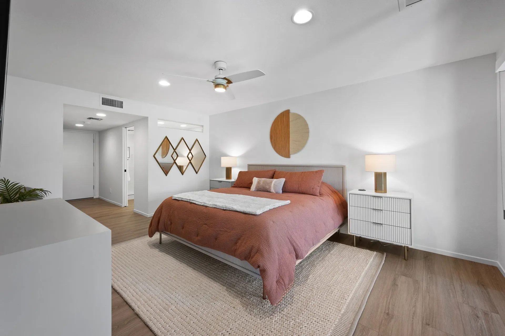 Bedroom with a large bed, rust-colored bedding, and two side tables with lamps. Wall decor includes circular art above the bed and diamond-shaped mirrors. Room has wooden flooring.