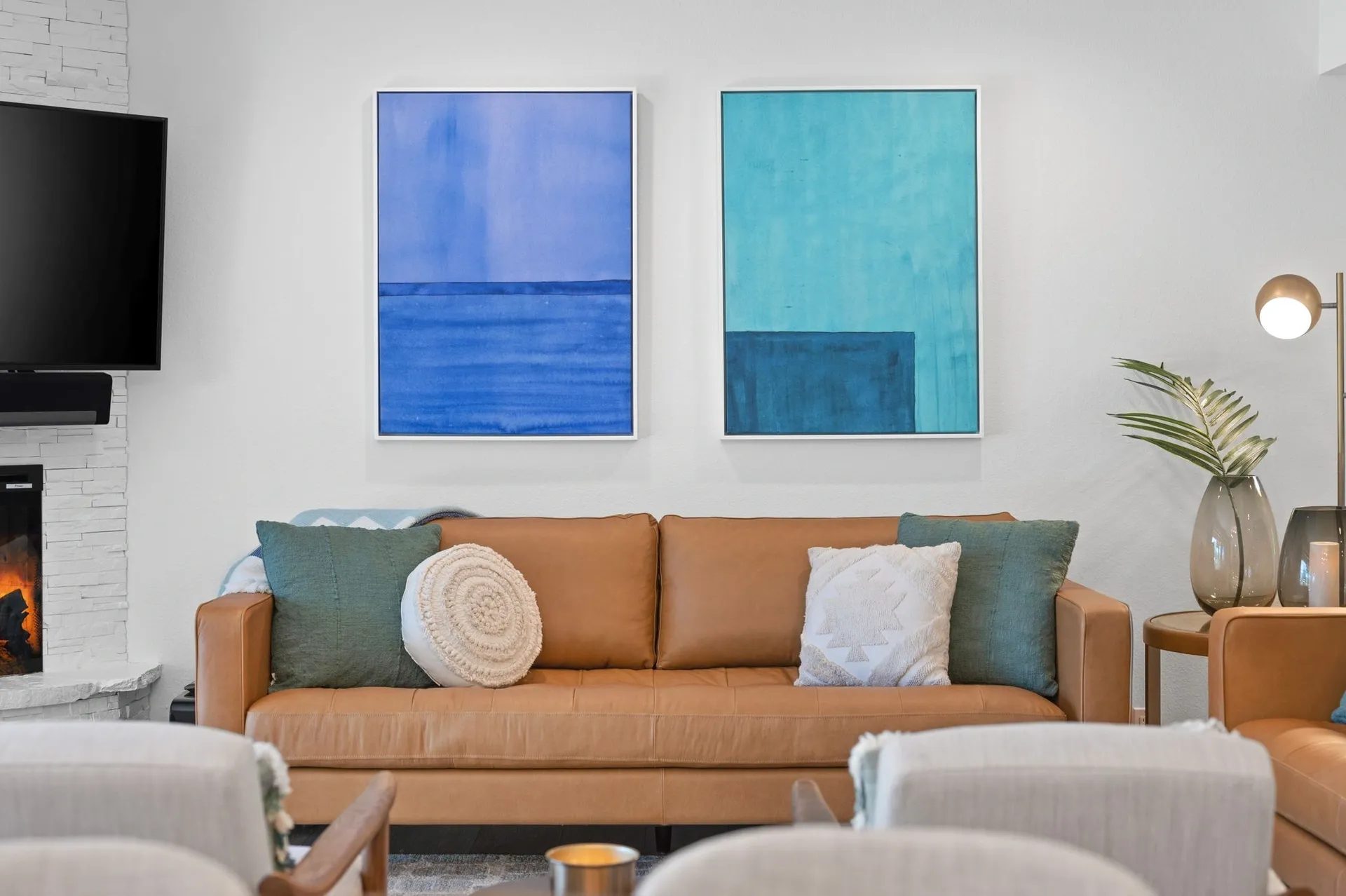 Living room with a brown leather sofa, abstract blue paintings on the wall, and a fireplace. Two armchairs are in the foreground, with a floor lamp and potted plant nearby.