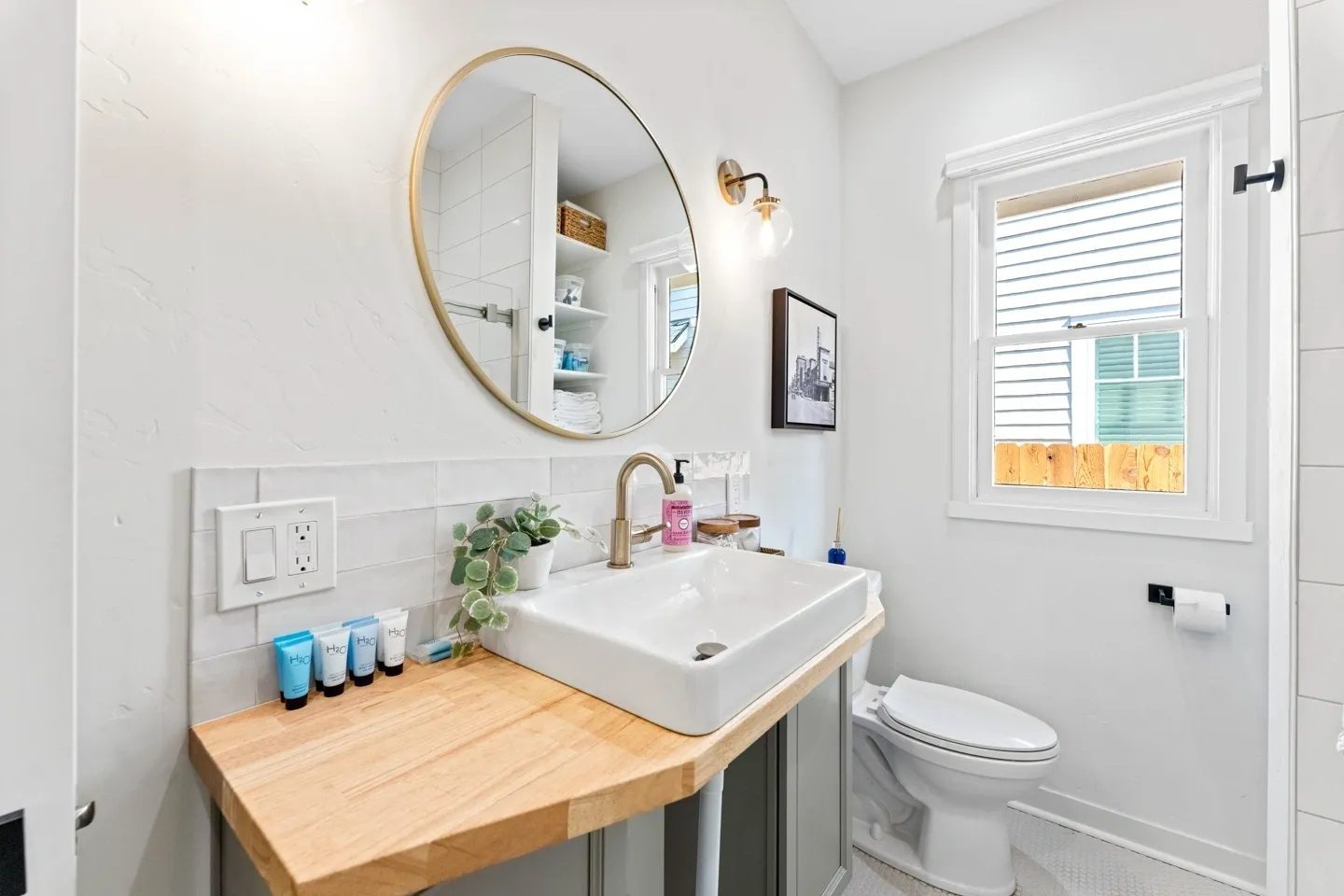 A bathroom features a round mirror, a sink with toiletries, and a small potted plant. There's a wall-mounted light, a window, and a toilet beside a wooden shelf.