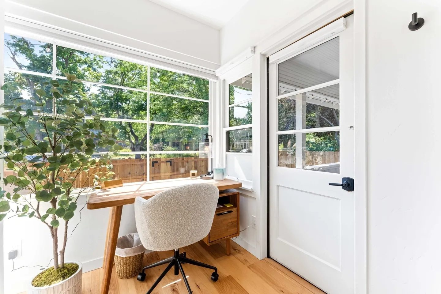 A bright home office with a desk, chair, lamp, and potted plant by a window. Sunlight streams through large panes, highlighting light wood flooring and a sliding door with a black handle.