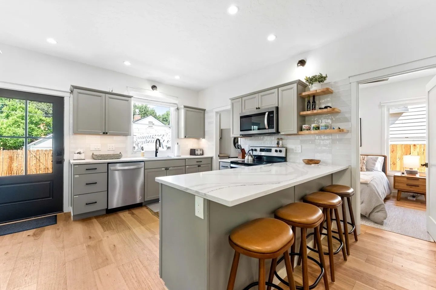 Modern kitchen with gray cabinets, white countertops, and stainless steel appliances. Wooden bar stools line the island. A door and window provide natural light. Bedroom visible in background.