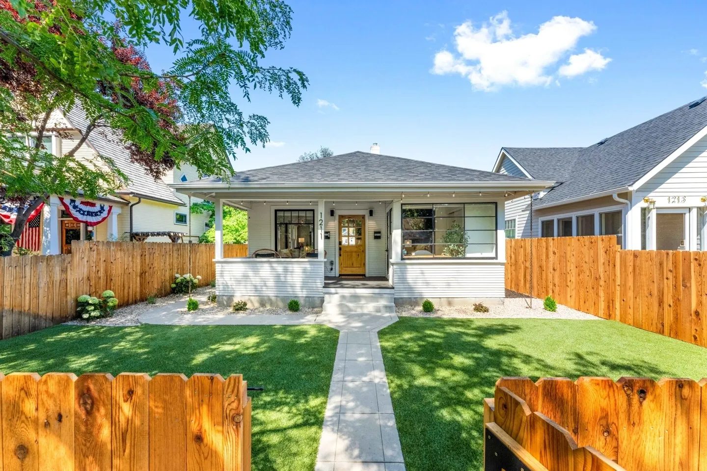 Single-story white house with a wooden front door and porch, surrounded by a wooden fence. A concrete walkway leads through a lawn. Neighboring houses are visible on both sides.