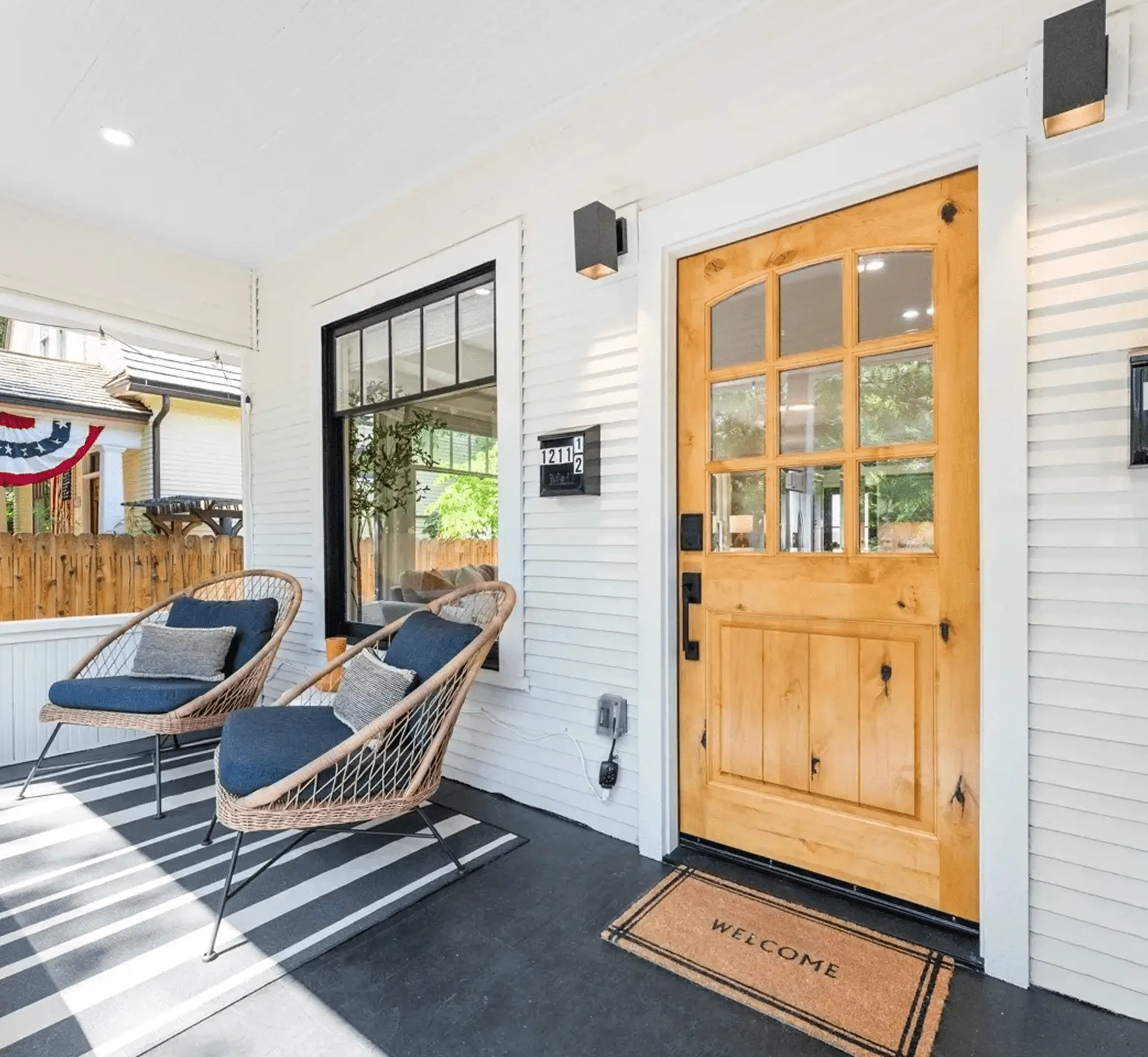 Front porch with a wooden door and welcome mat, two wicker chairs with cushions, black and white striped rug, and U.S. flag decor on a nearby railing.